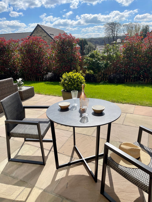 A bistro round table made of metal with a laminate top, presented in a outdoor setting with a black matte finish, accompanied by two chairs, set in a garden environment.